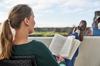 Eine Frau liest auf dem Balkon mit hochgelegten Beinen einen Nordsee-Krimi bei Sonnenschein.
